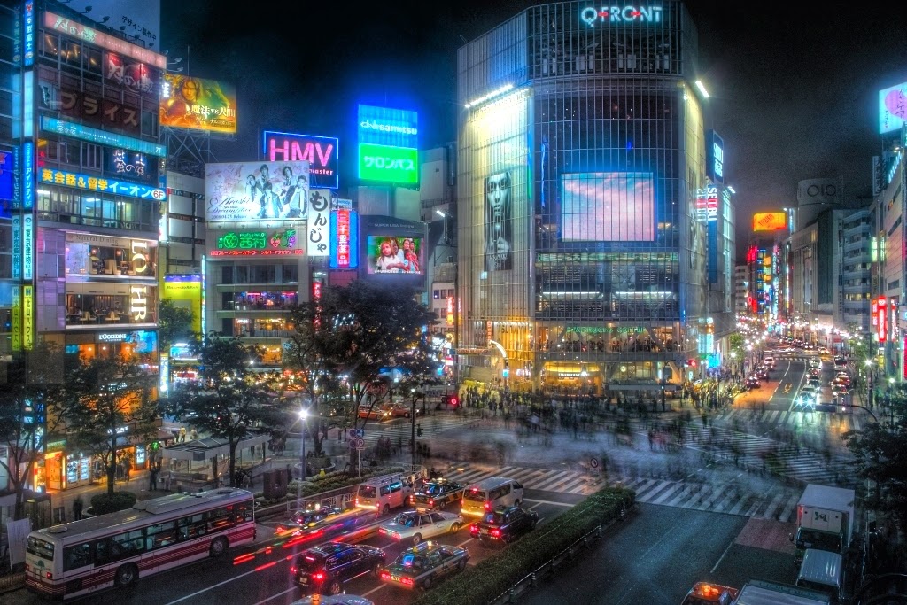 Shibuya at night — neon signs, crossing crowds, and taxi lights, photographed from above.